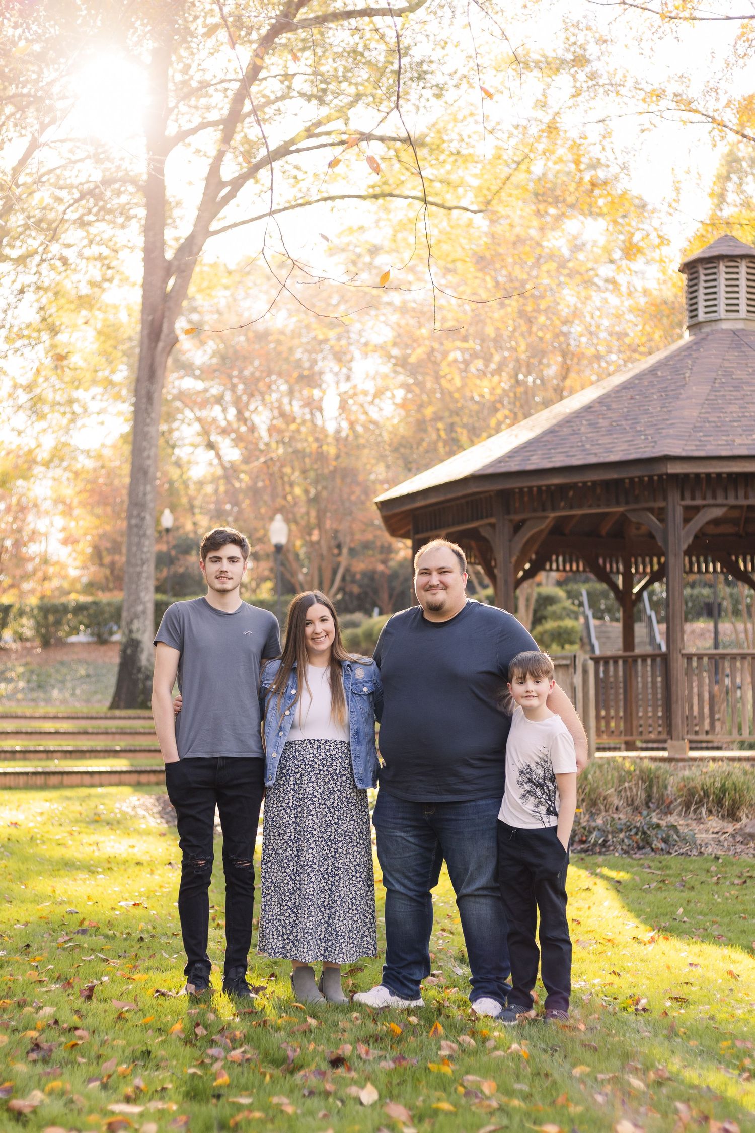 A group poses outdoors near a gazebo surrounded by fall foliage and dappled sunlight on grass.