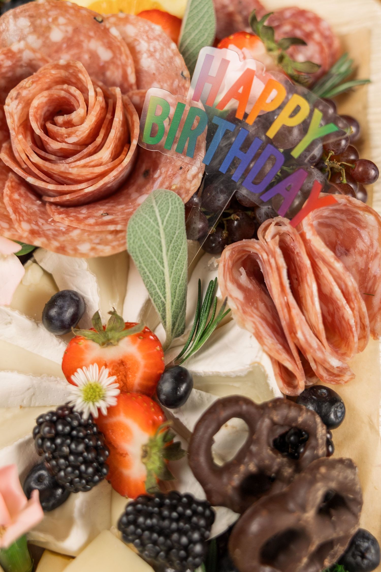 Gourmet charcuterie board decorated with rainbow Happy Birthday sign, meats, berries and chocolate pretzels.