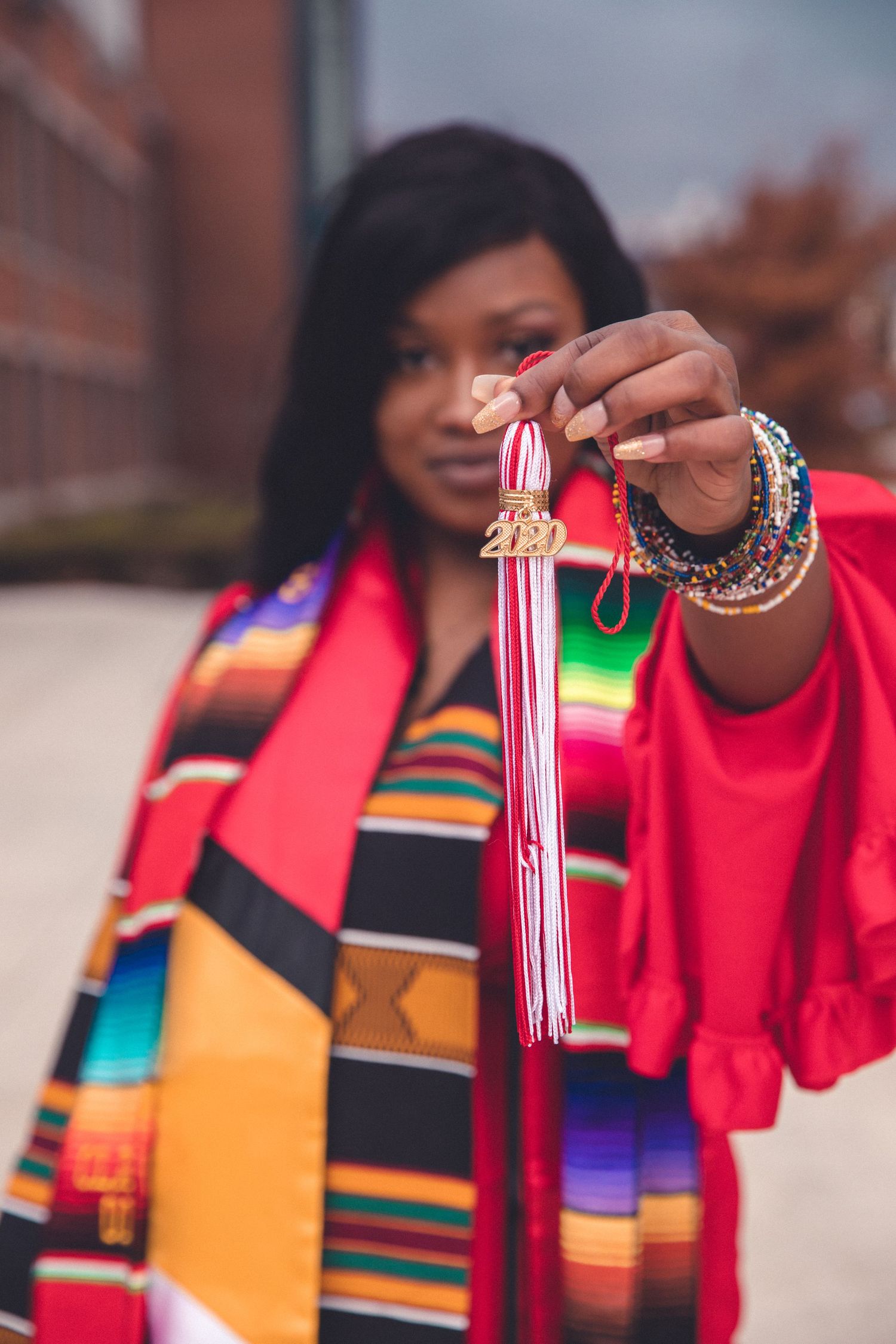 Graduate holding tassel wearing colorful kente stole and red graduation gown outdoors.