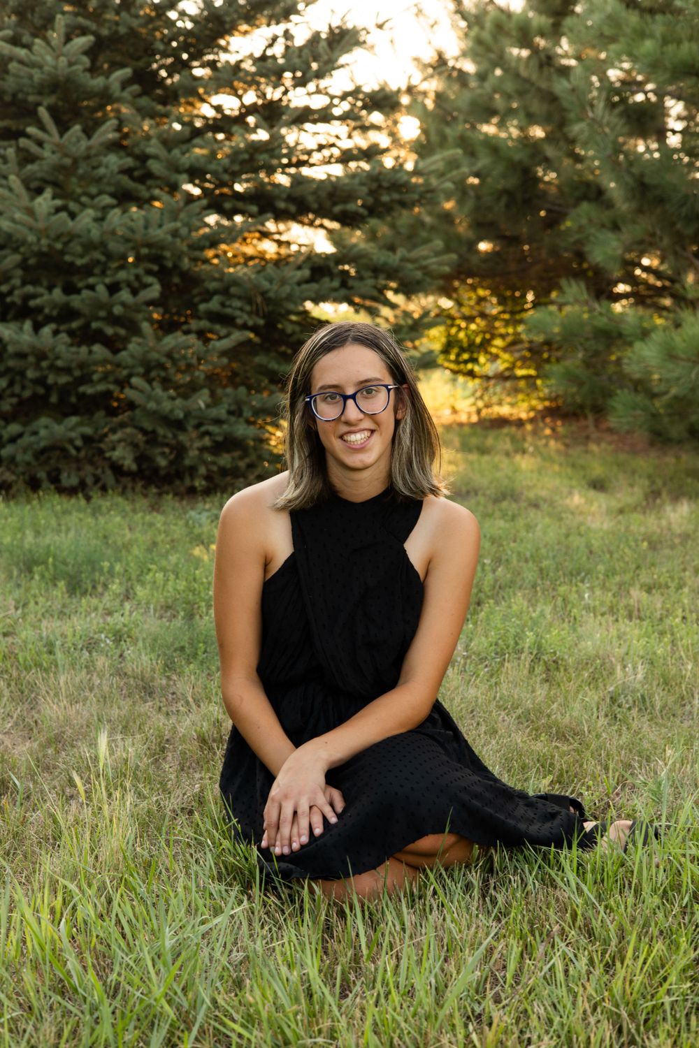 Portrait of someone in a black sleeveless dress sitting in grass at sunset with evergreen trees in background.