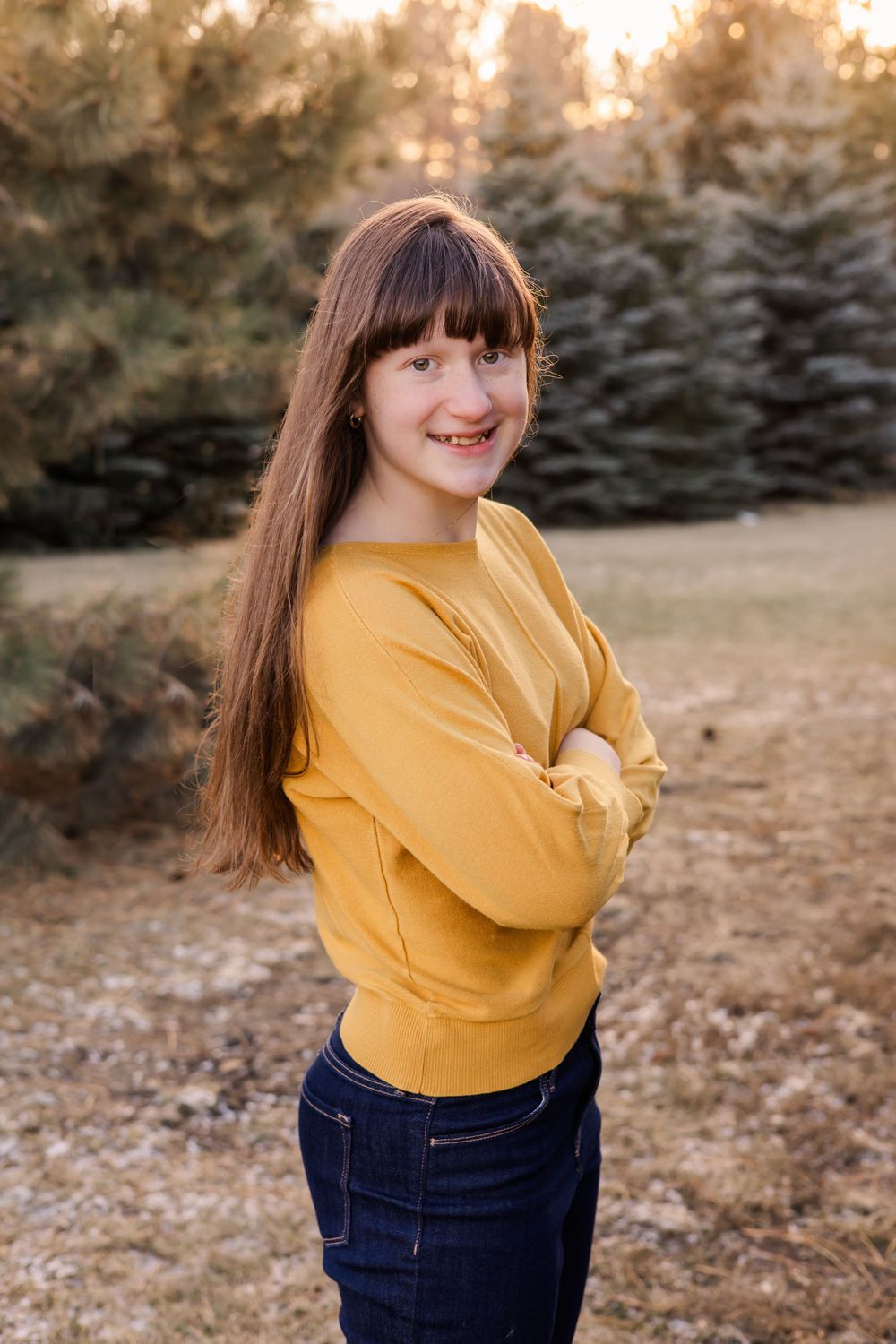 Outdoor portrait in yellow sweater and jeans posed against evergreen trees at golden hour.