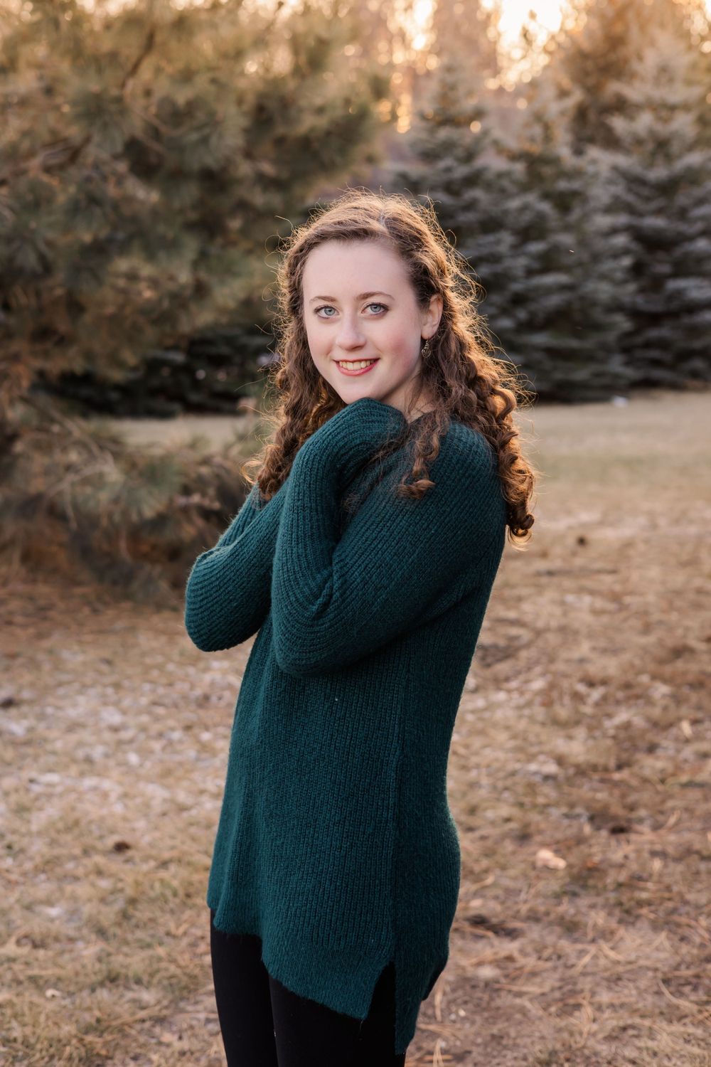Outdoor portrait in dark green sweater posed against evergreen trees at sunset.