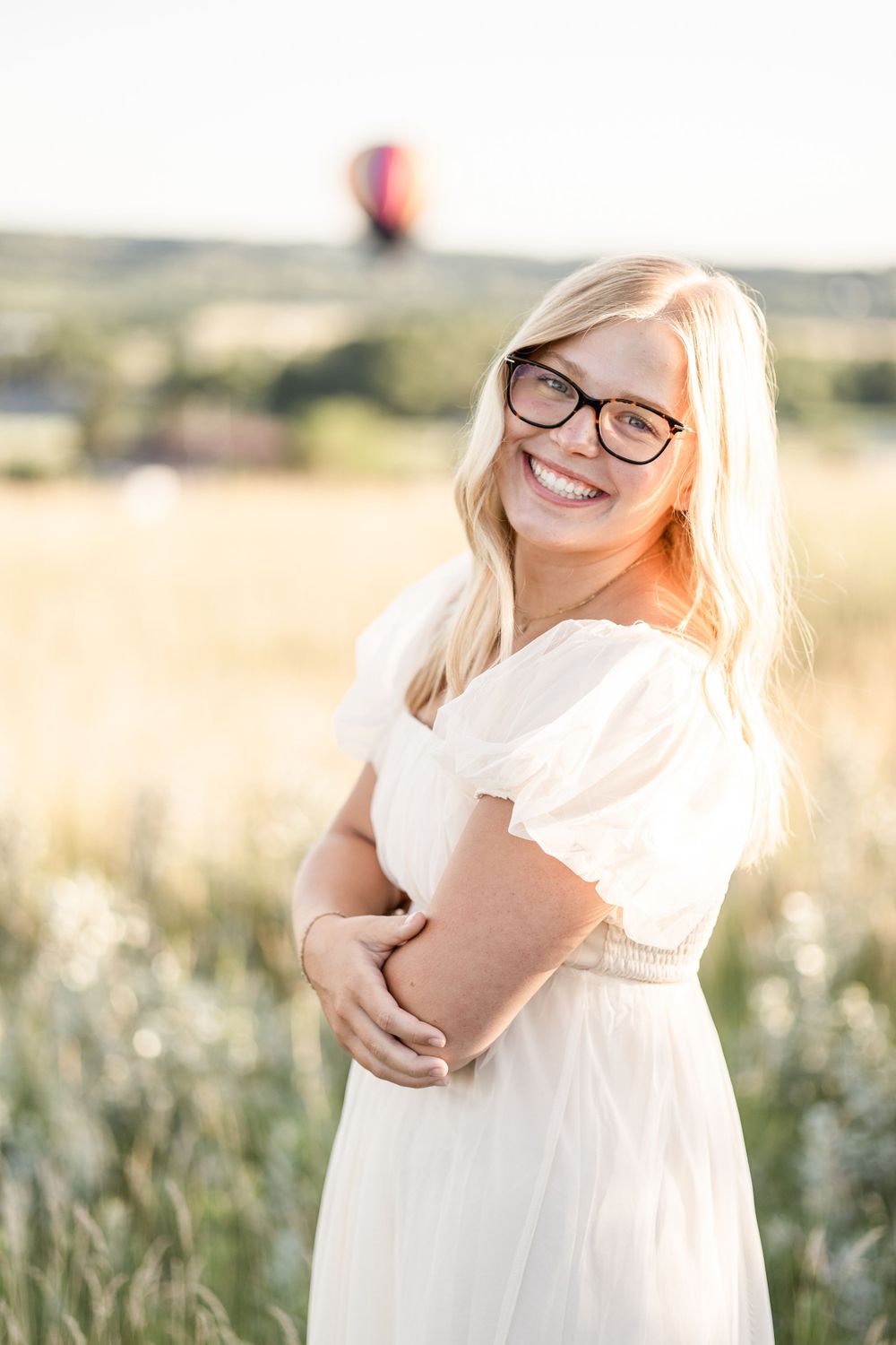 Person in white dress smiling outdoors with sunlit field in background.