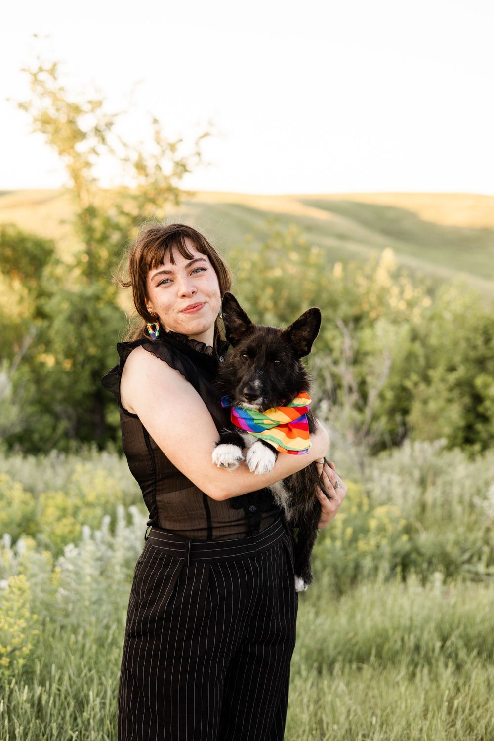 A woman holds a black dog wearing a rainbow bandana in a sunny meadow.