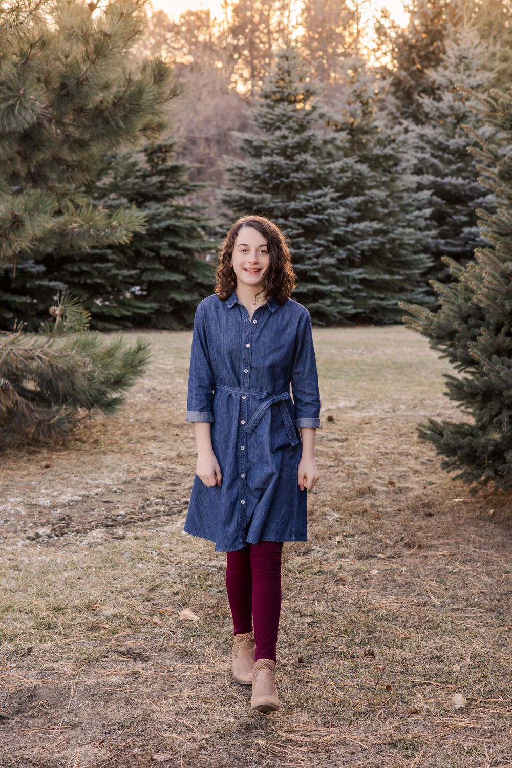 Portrait in denim dress and burgundy leggings walking among evergreen trees at sunset.
