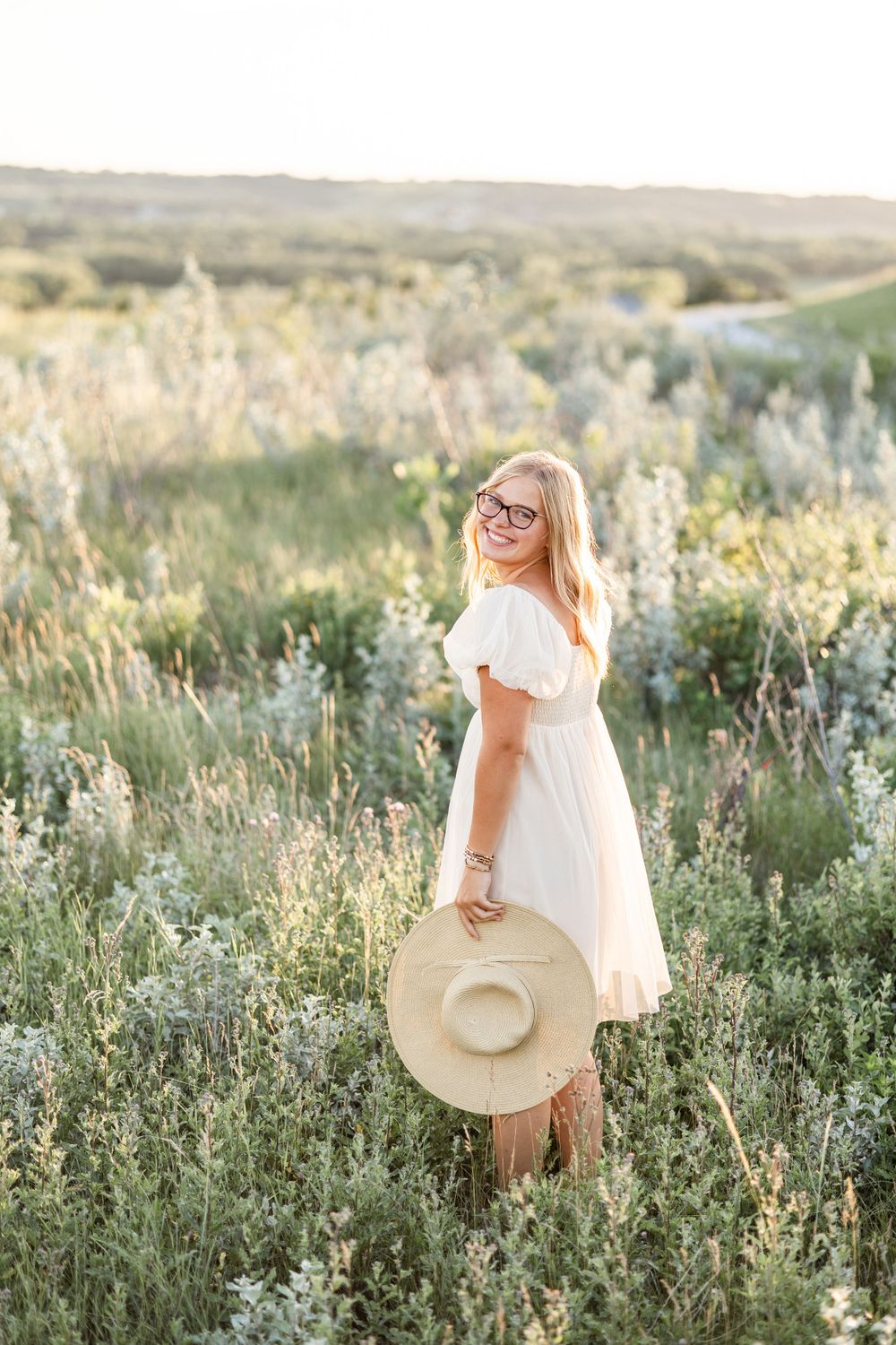 Person in flowing white dress holding sun hat in wildflower meadow.