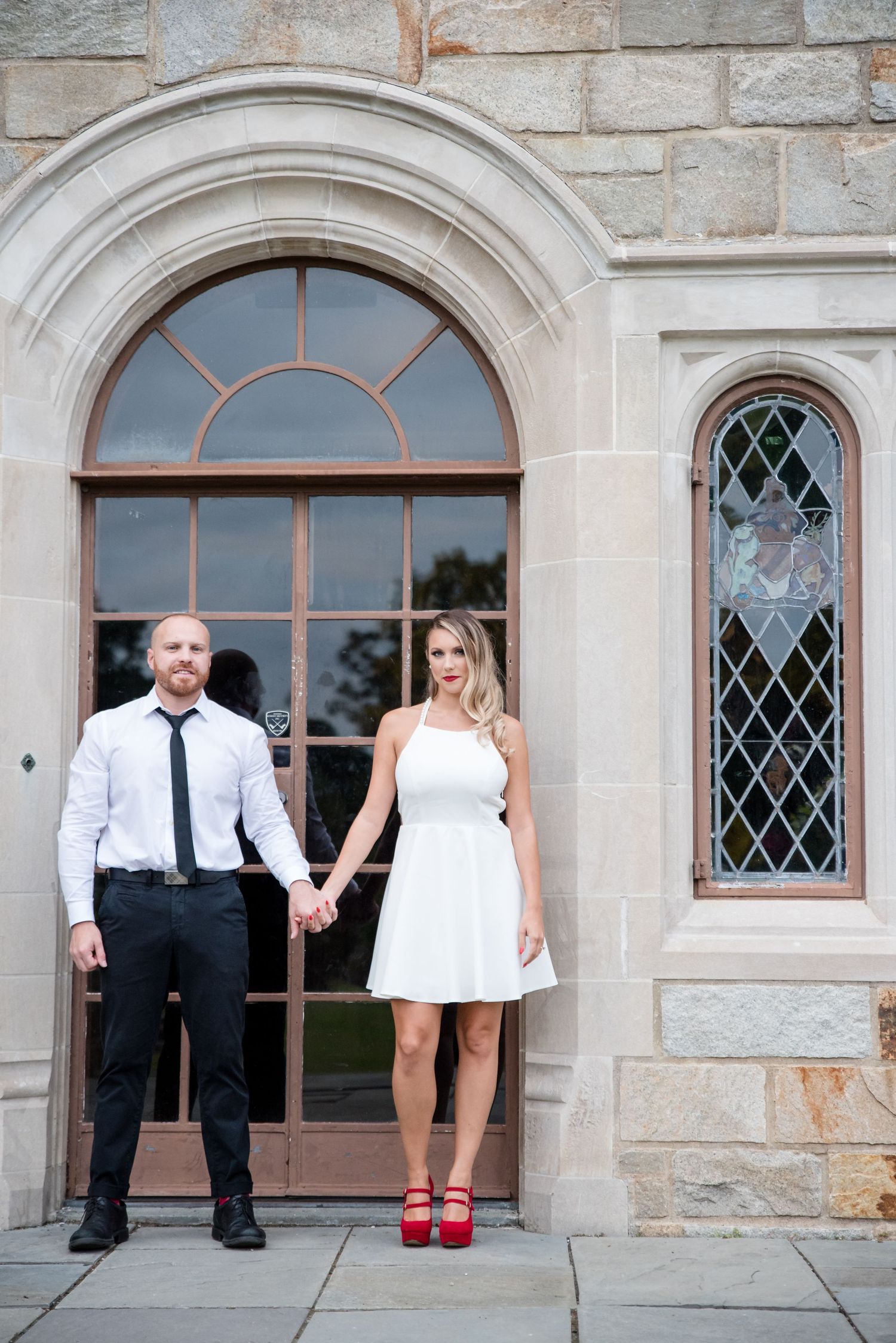 Bride in a white mini dress and red shoes poses with groom in front of stone church entrance with arched windows.
