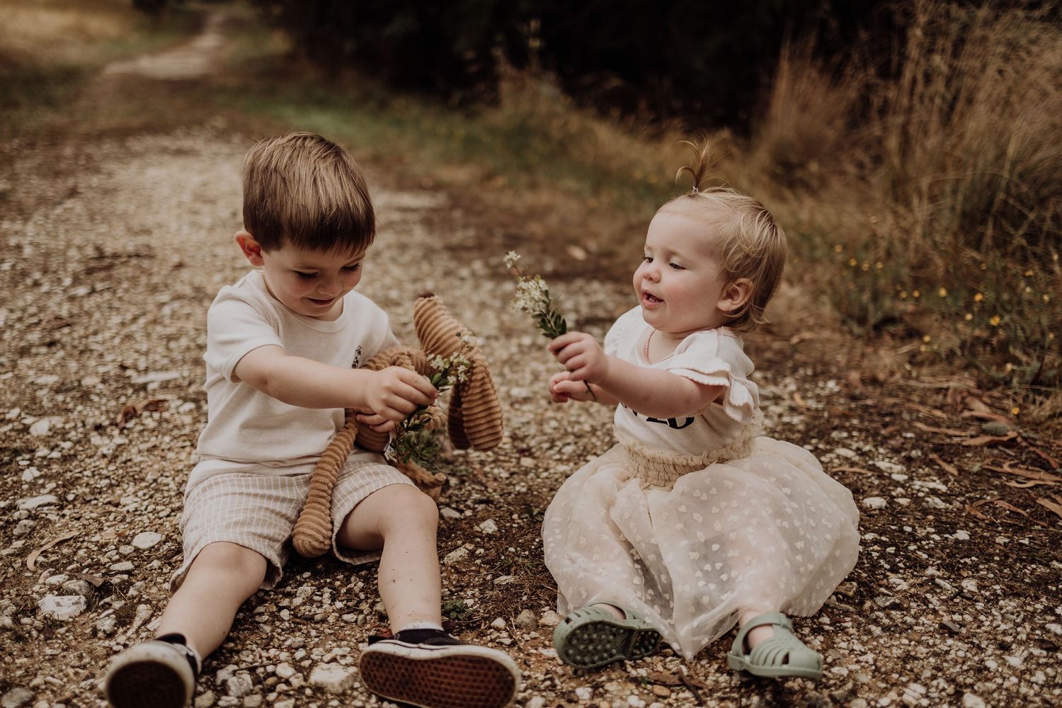 Two small children in light colored outfits play together on a dirt path.