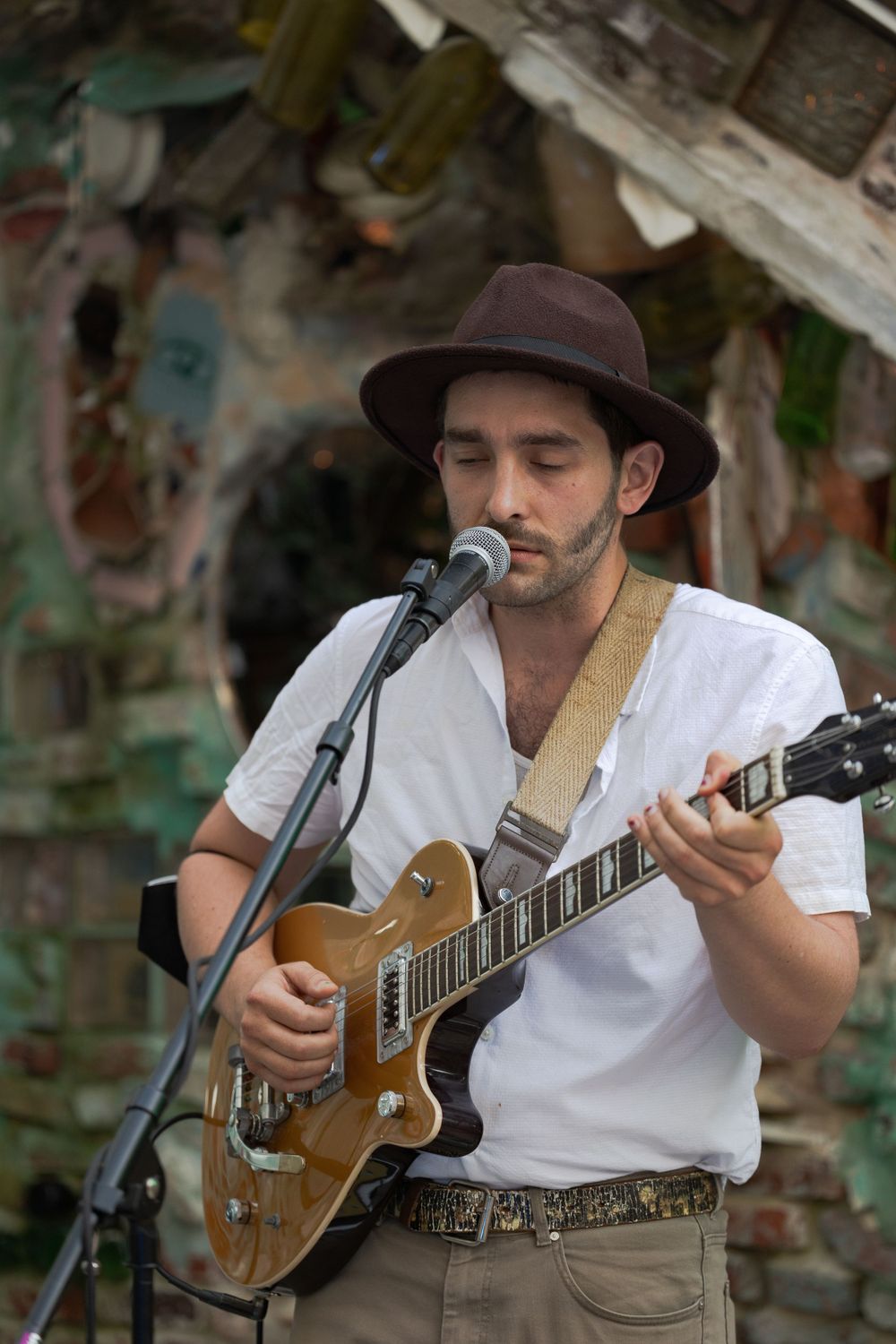 Folk musician in brown hat playing acoustic guitar and singing into microphone outdoors.