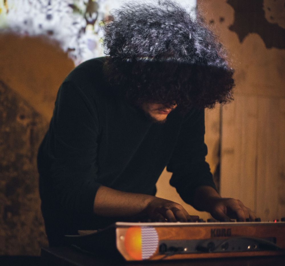 Musician with curly hair playing keyboard in dim lighting with orange glow.