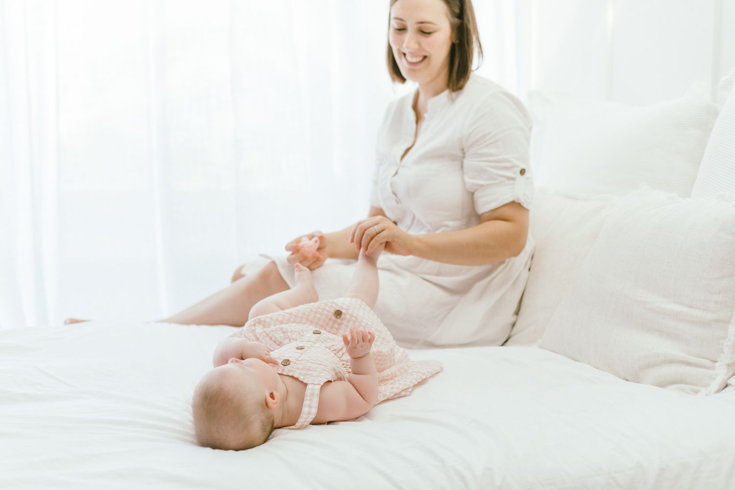 Mother and baby share intimate bonding moment on white bedding in natural light.