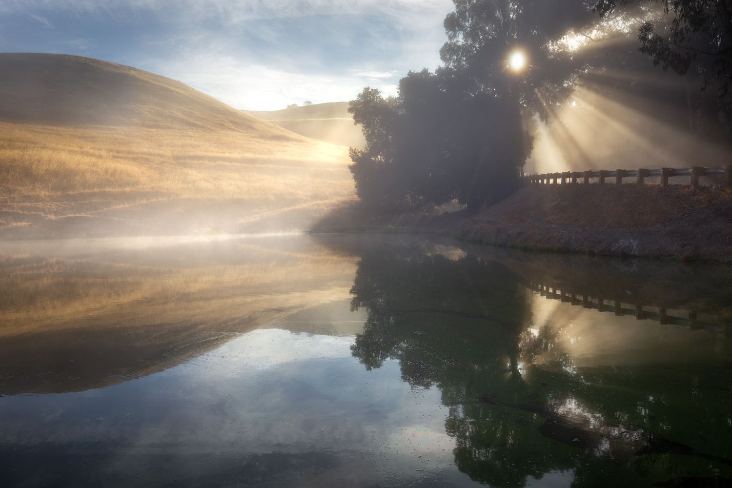 Sunbeams pierce through morning mist over a tranquil lake with trees and hills reflected in the still water.