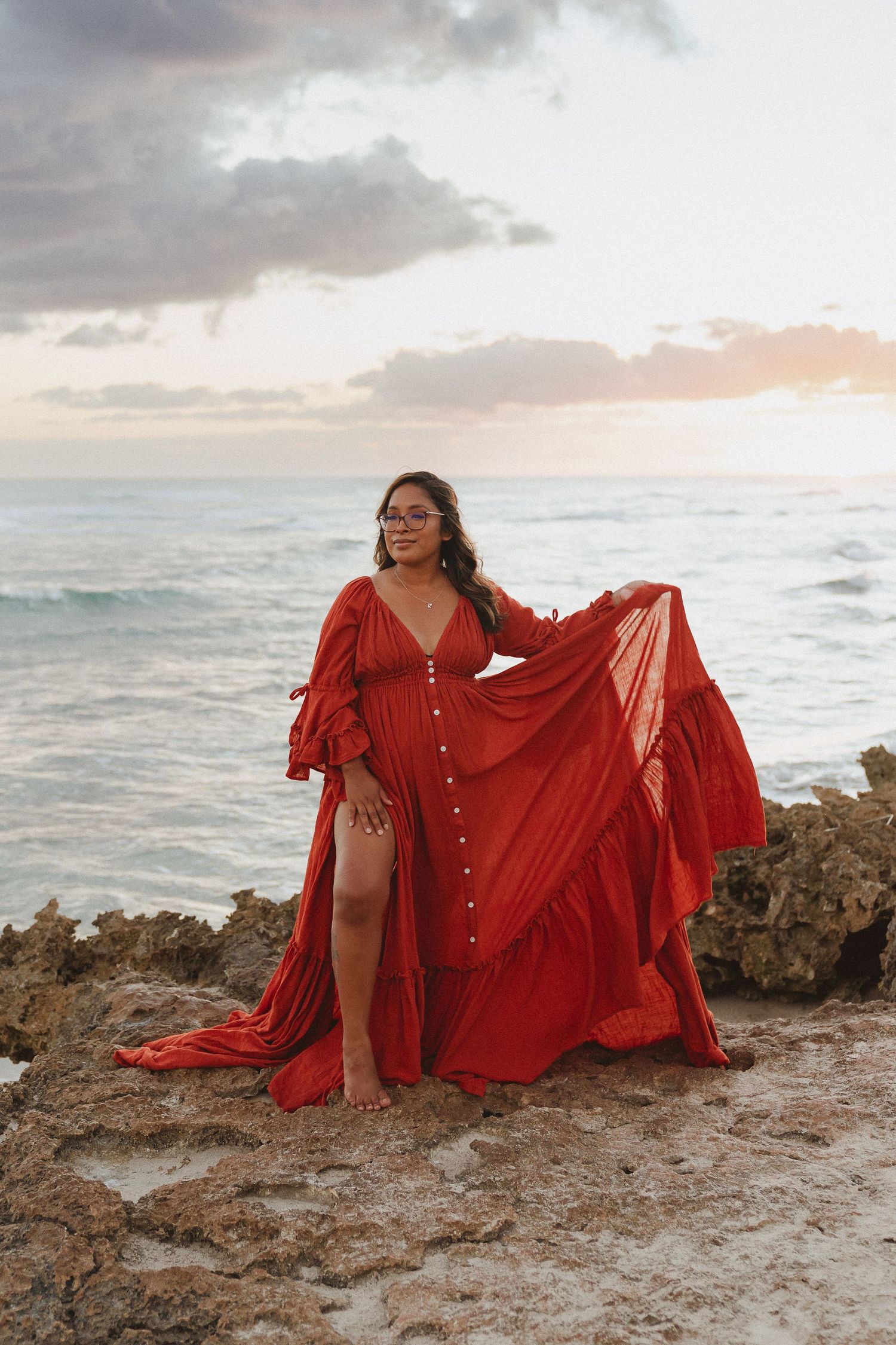 Photographer Kelly Pagan of Moments With Kell on the rocks by the ocean with her burnt orange dress on Oahu in Hawaii