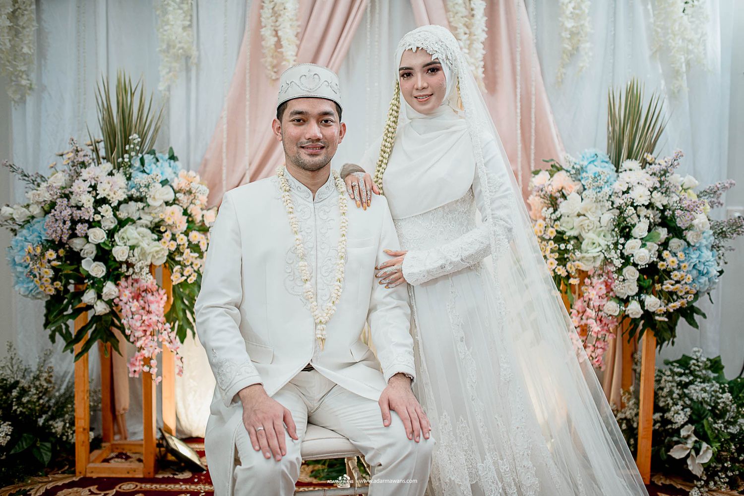 Wedding portrait of couple in matching white Islamic attire with floral decorations.