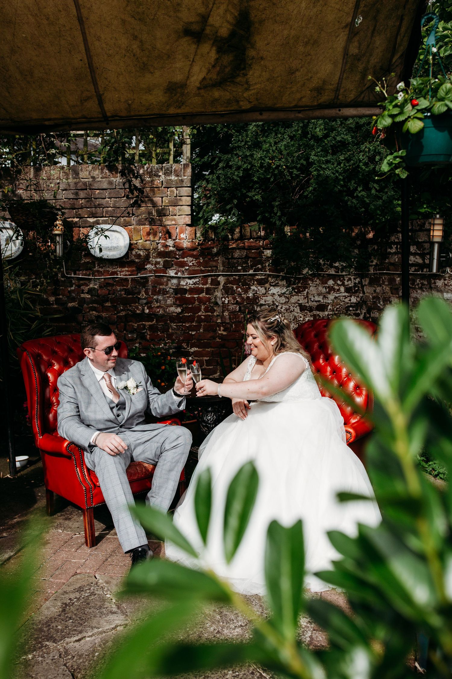 Couple sharing drinks on red vintage chairs against rustic brick wall with greenery.