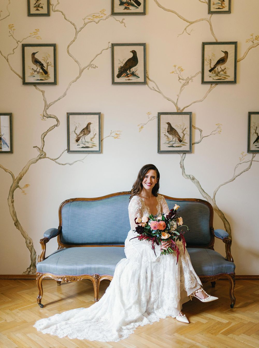 Bride sitting on blue couch with bird-themed wallpaper in Lobkowicz Palace, Prague.