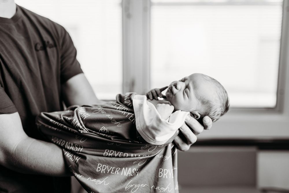 Father cradling a swaddled newborn baby in his arms in a hospital room, black and white photo.