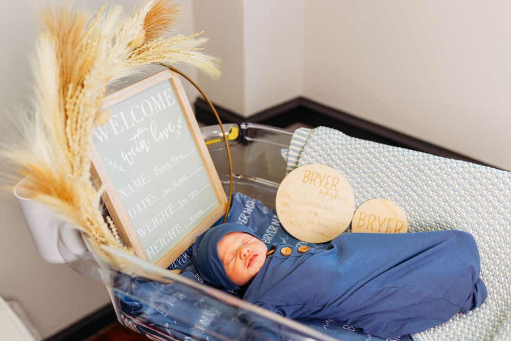 Newborn baby in blue swaddle sleeps in bassinet with wooden milestone discs and welcome sign.
