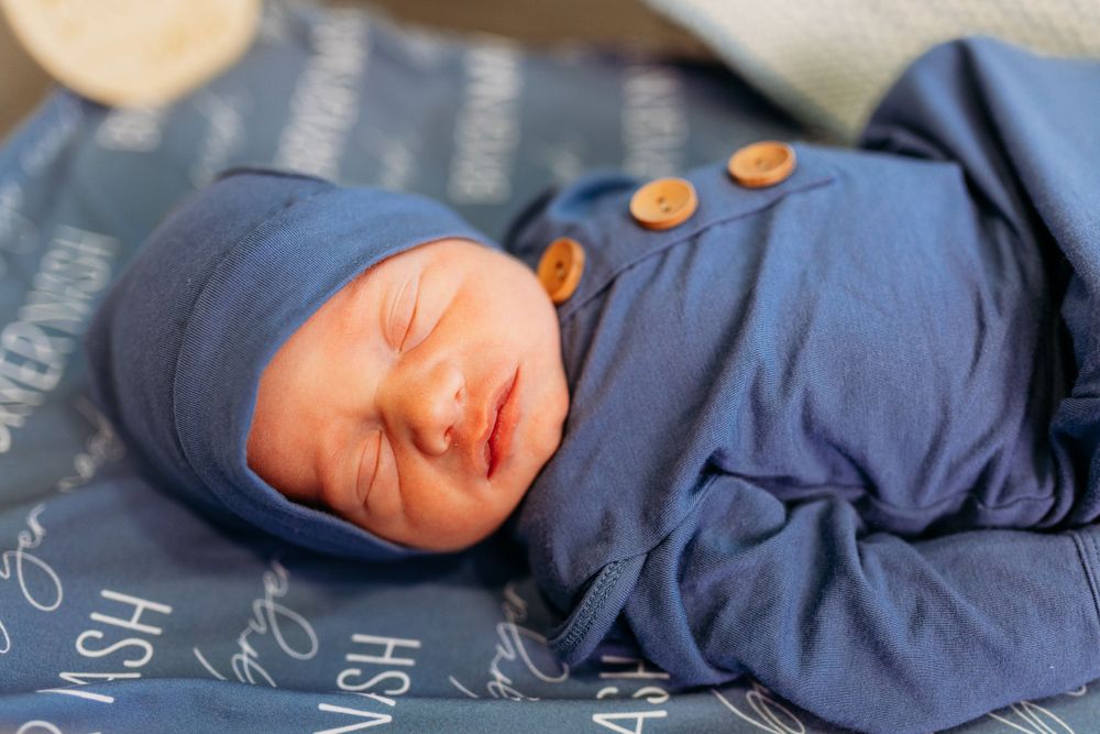 Close-up of sleeping newborn wearing a blue hat and outfit with decorative wooden buttons.