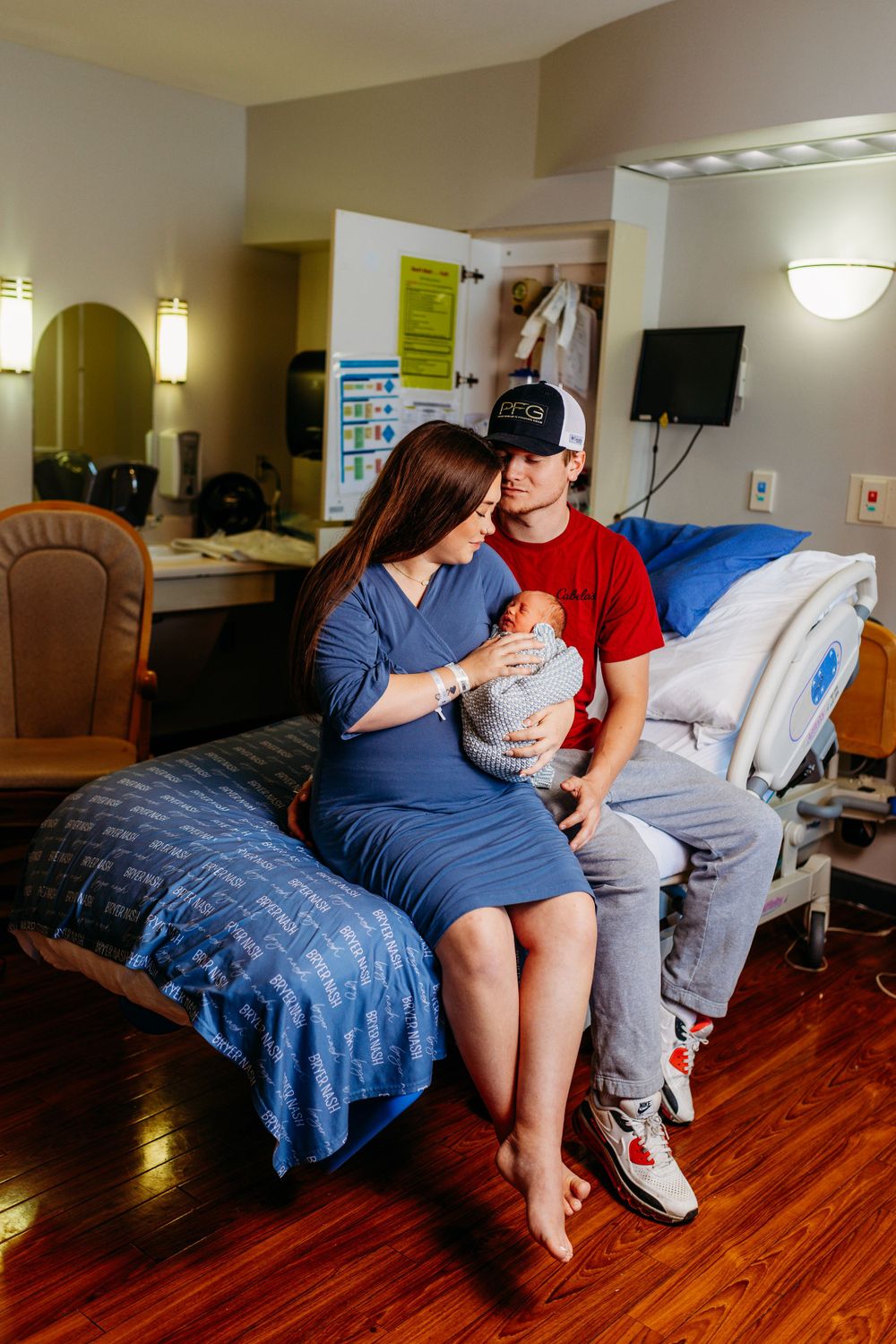 New parents sit on hospital bed admiring their swaddled newborn baby together.