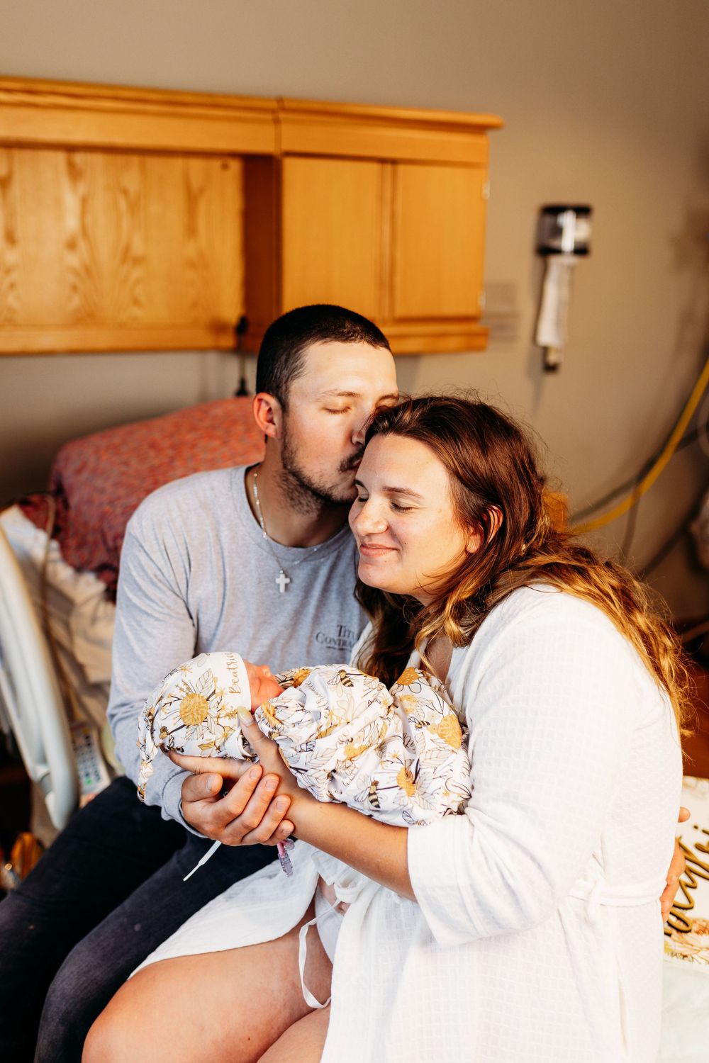 New parents share a tender moment as father kisses mother's head while she holds their swaddled newborn in hospital room.