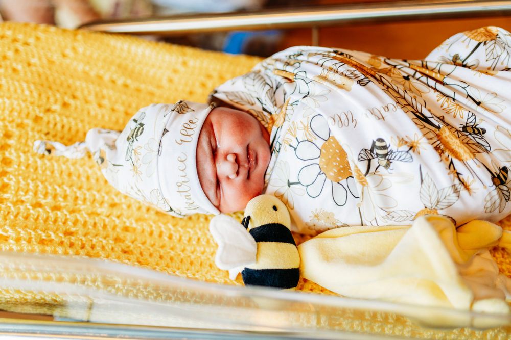 Newborn baby swaddled in a floral wrap lying on a yellow knit blanket with a bee plush toy.