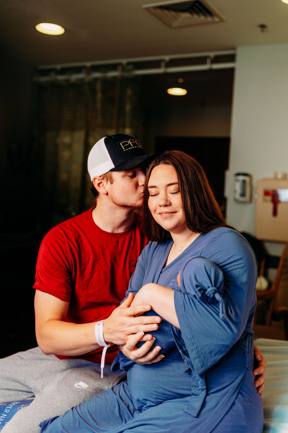 Expectant father kisses pregnant mother tenderly on cheek in hospital room.