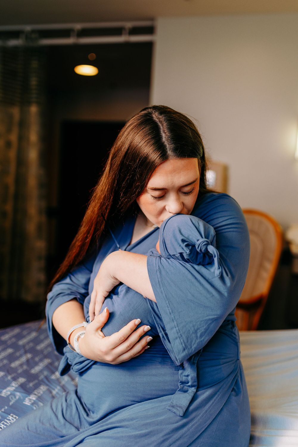 New mother in blue robe lovingly kisses her newborn baby on hospital bed.