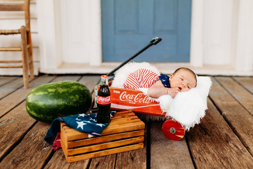 Newborn baby in patriotic outfit sleeping in red Coca-Cola wagon with watermelon and American flag.