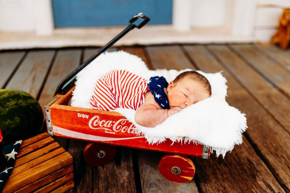 Newborn baby in striped outfit sleeping on white fur in vintage red Coca-Cola wagon outdoors.