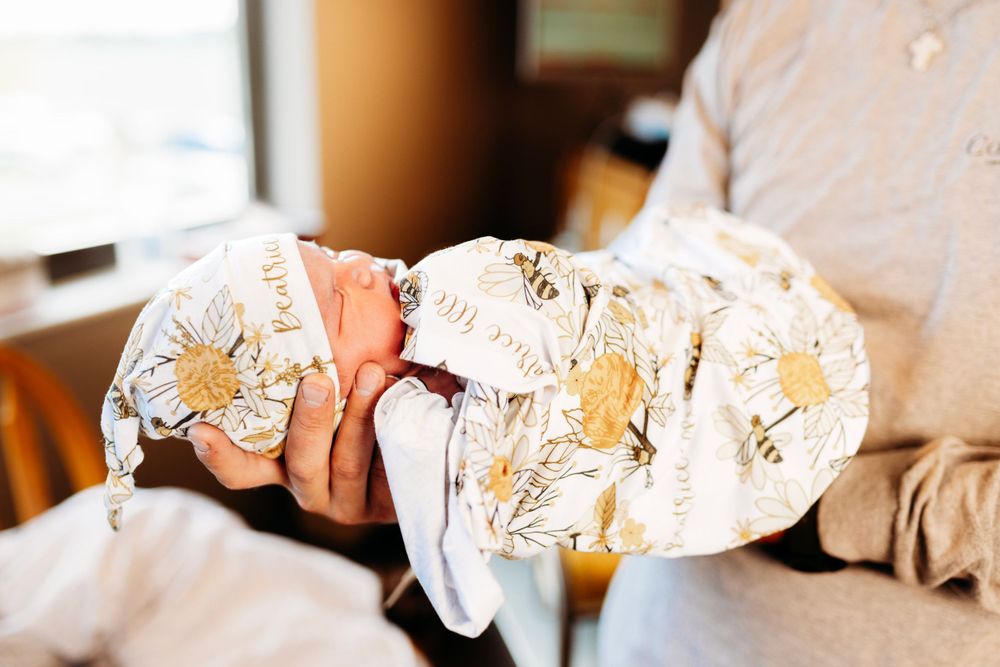 Close-up of father's hands carefully cradling tiny swaddled newborn baby in floral blanket near hospital window.