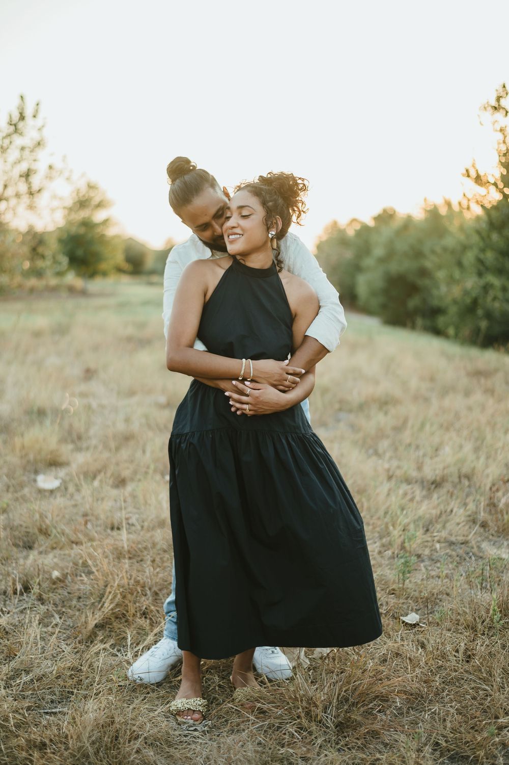 Intimate embrace in a sunlit meadow with black halter dress and casual white top.