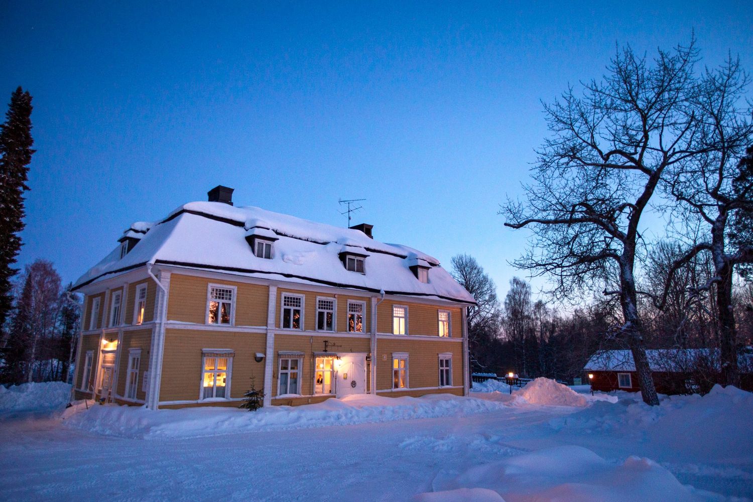 Snow-covered yellow manor house glowing warmly at twilight surrounded by deep winter snow.