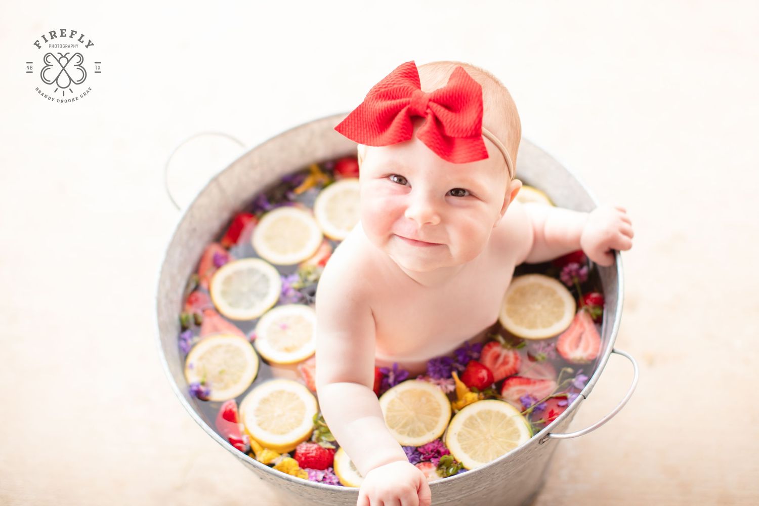 Baby in red bow sits in decorative bowl with citrus fruit slices.