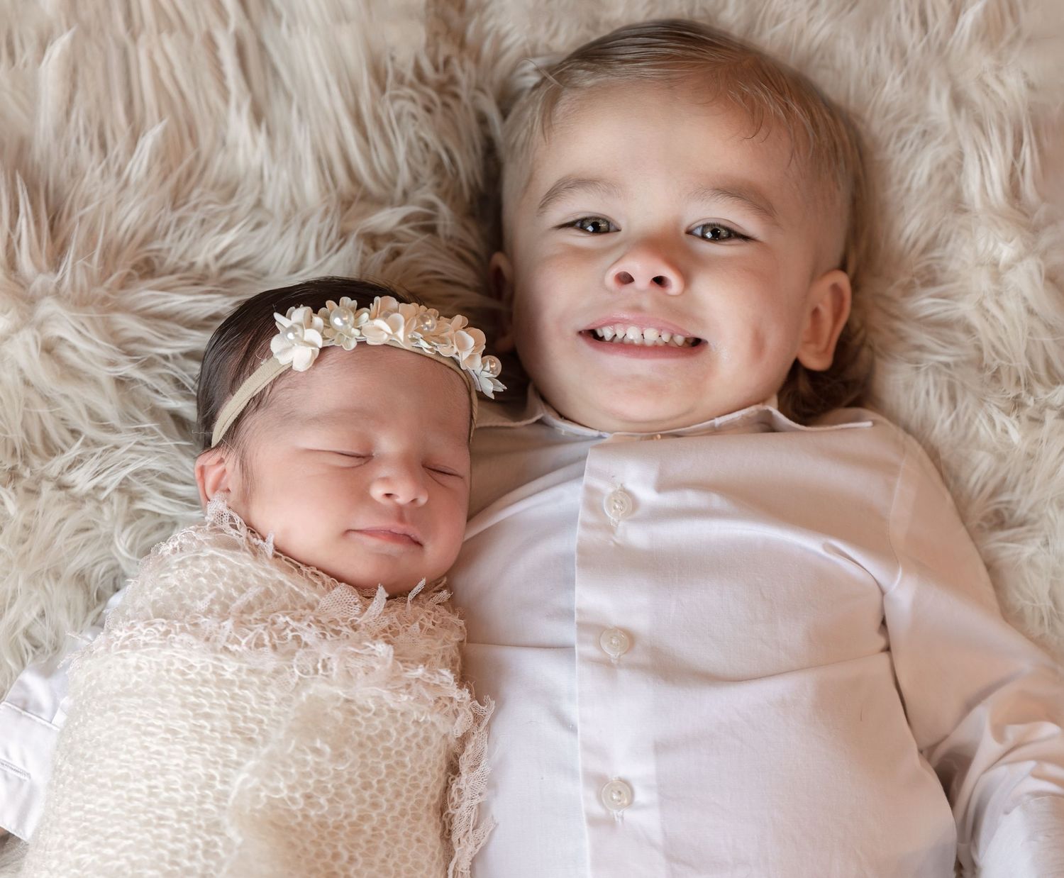 Two siblings lying on plush fur, one sleeping newborn and older sibling smiling at camera.