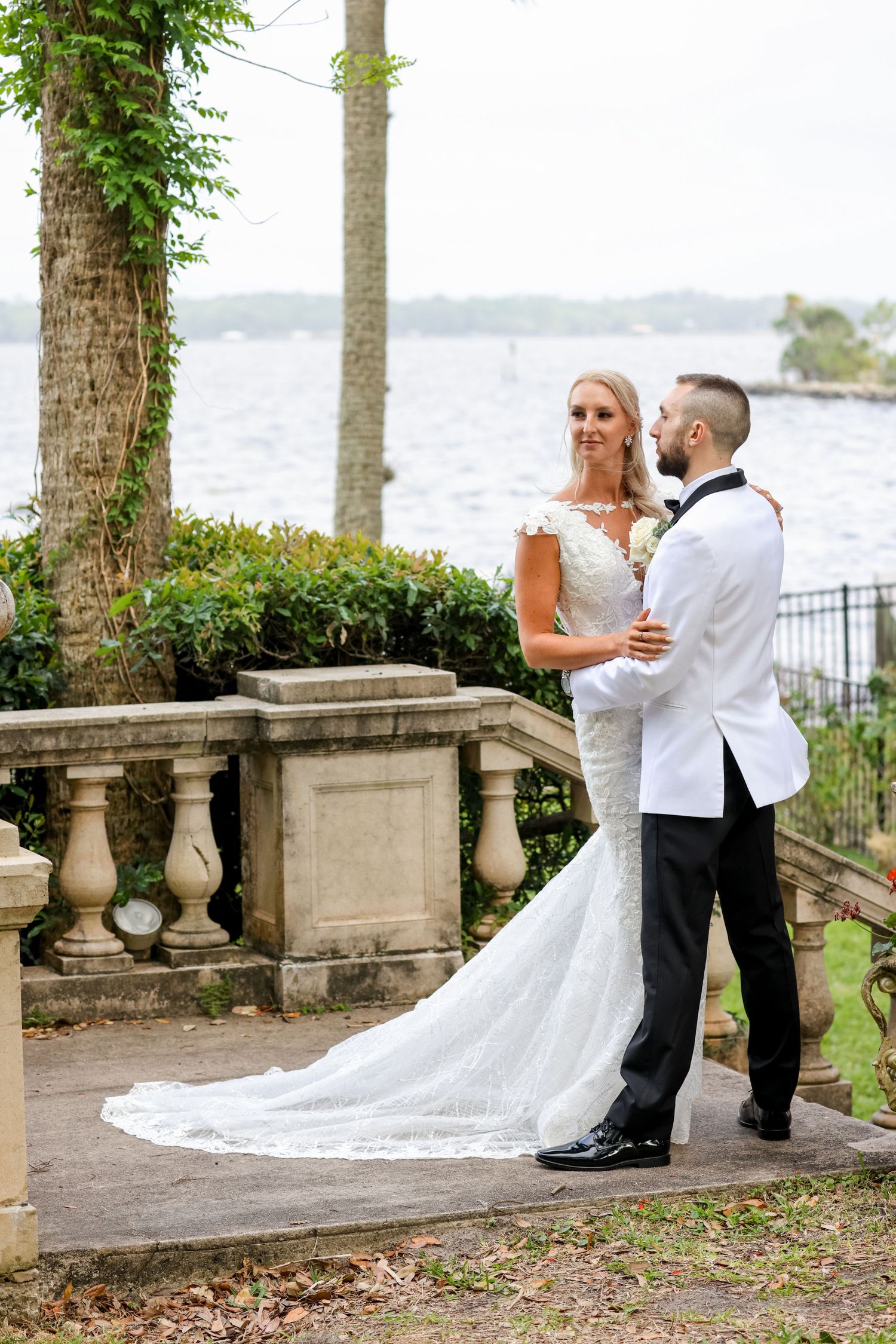 A couple shares an intimate moment on their wedding day along a waterfront terrace with stone balustrades and palm trees.