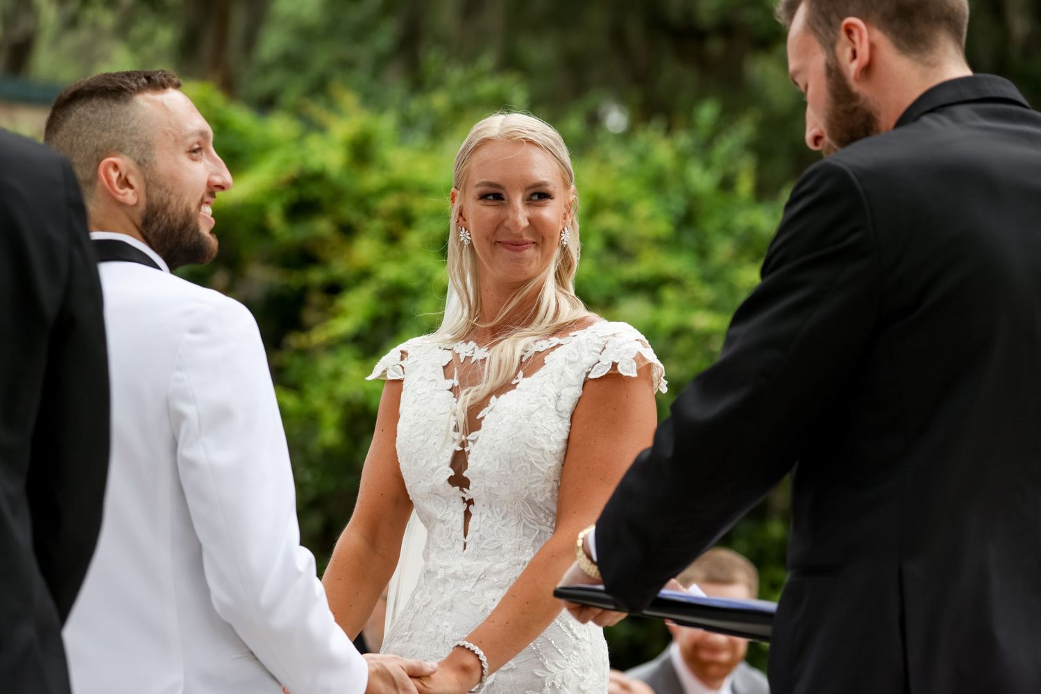 A bride in an elegant white lace wedding dress smiles during an outdoor ceremony with guests and officiant.