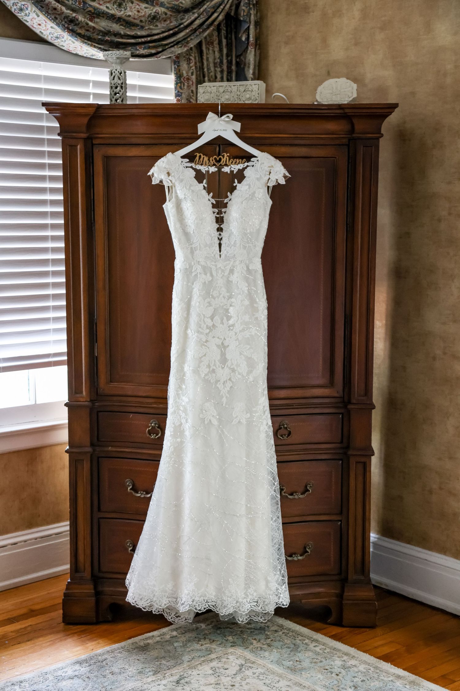 White lace wedding dress with plunging neckline hanging on wooden hanger in front of dark wood armoire.