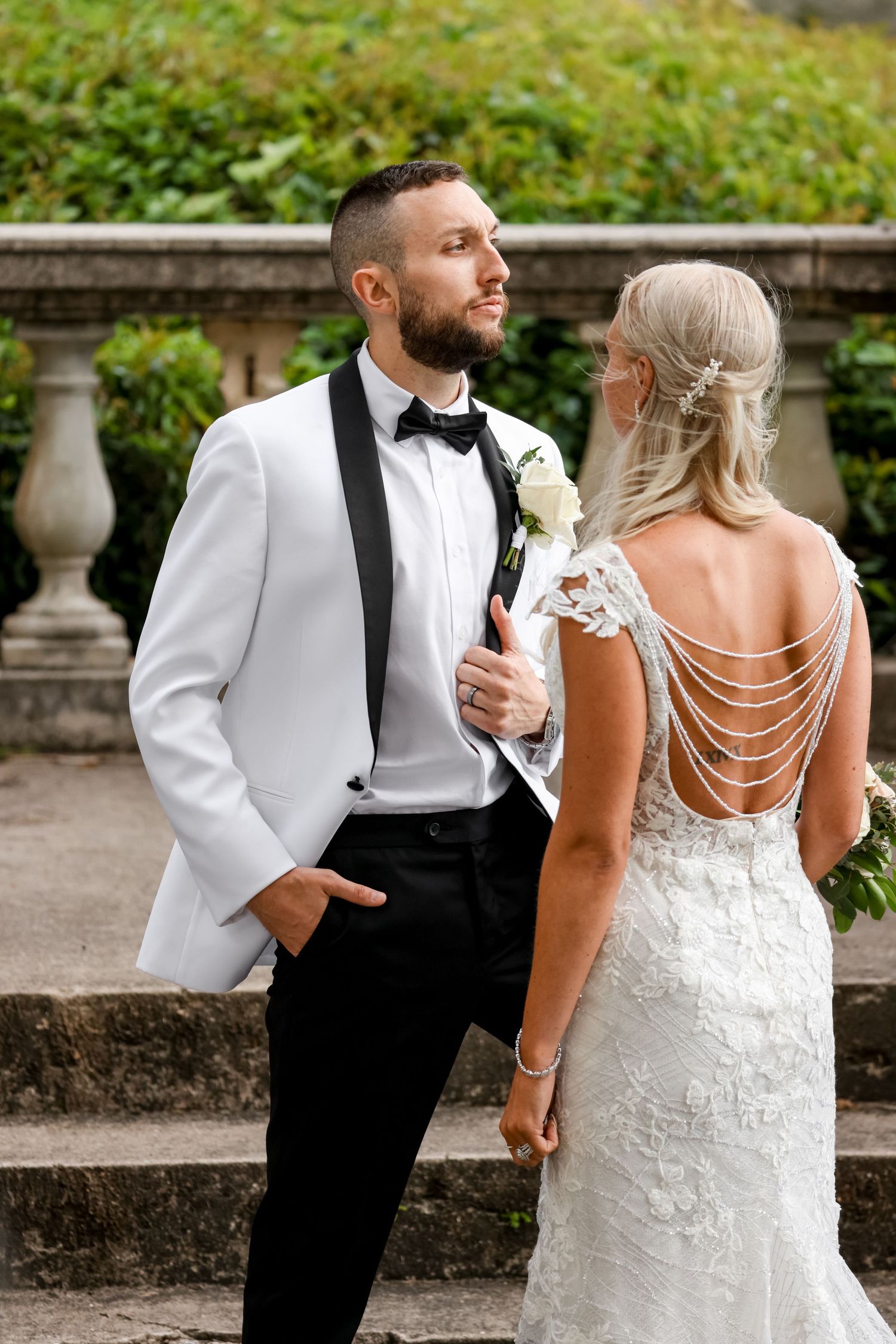 A bride in a backless white wedding gown with pearl draping and groom in white tuxedo jacket stand on stone steps.