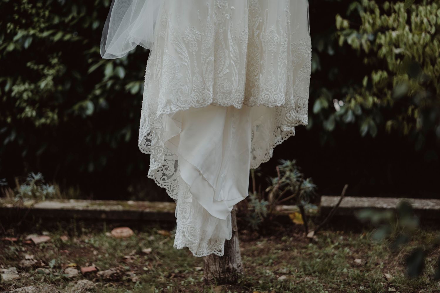 A delicate white lace wedding dress hanging outdoors against dark foliage.
