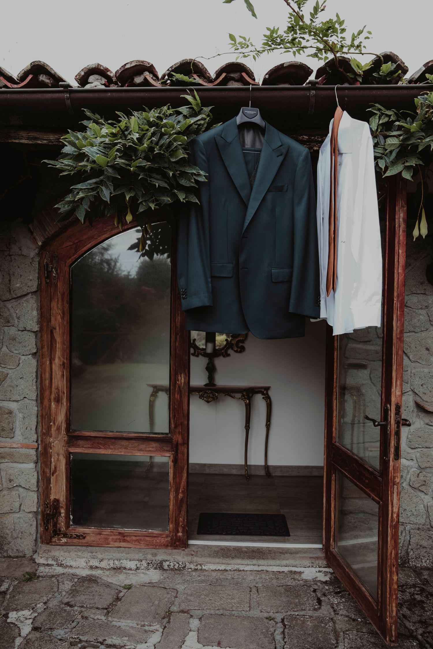 Navy suit jacket and white shirt hanging in rustic wooden doorframe with greenery overhead.