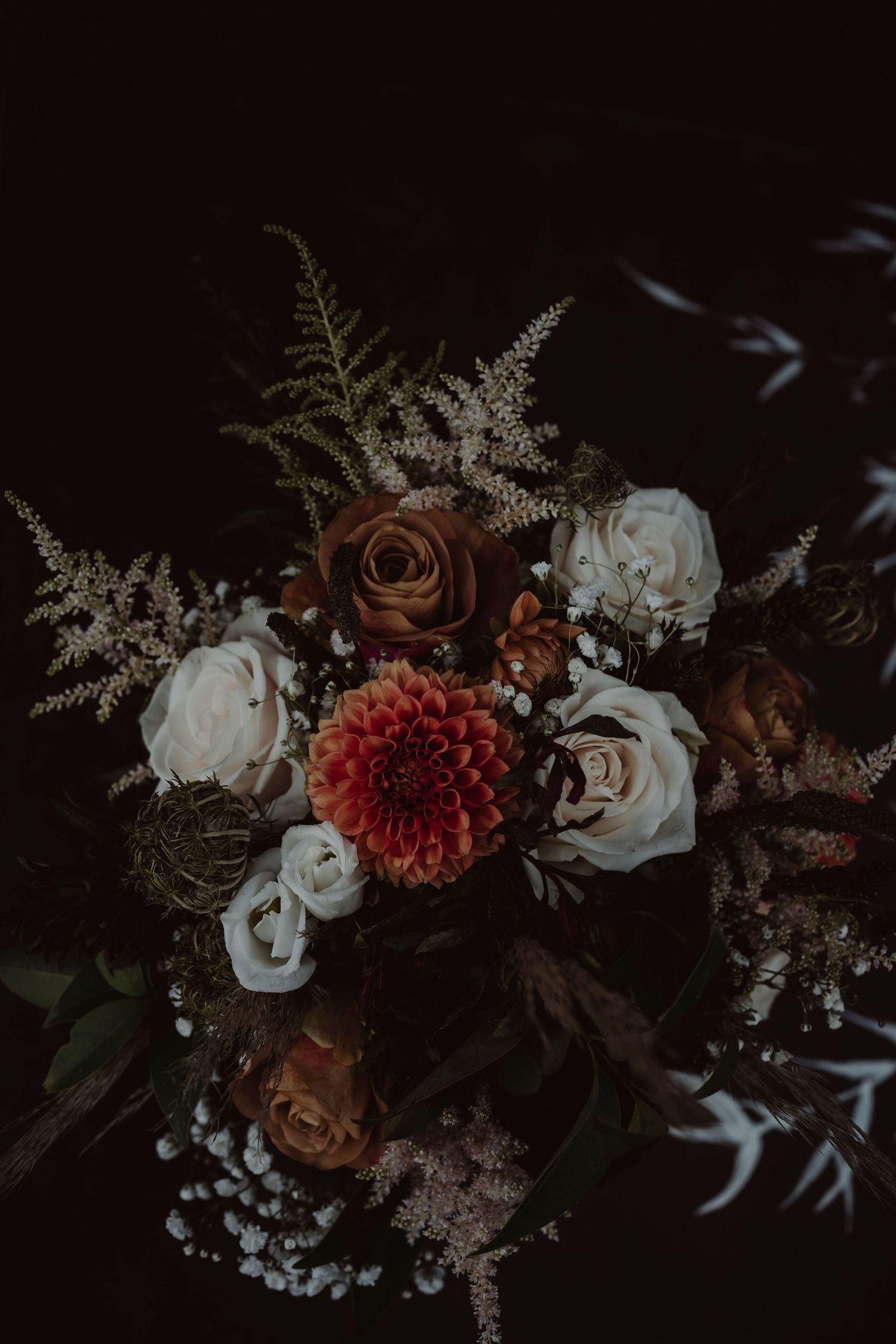 Moody floral arrangement featuring white roses, red dahlia, and delicate ferns against dark background.