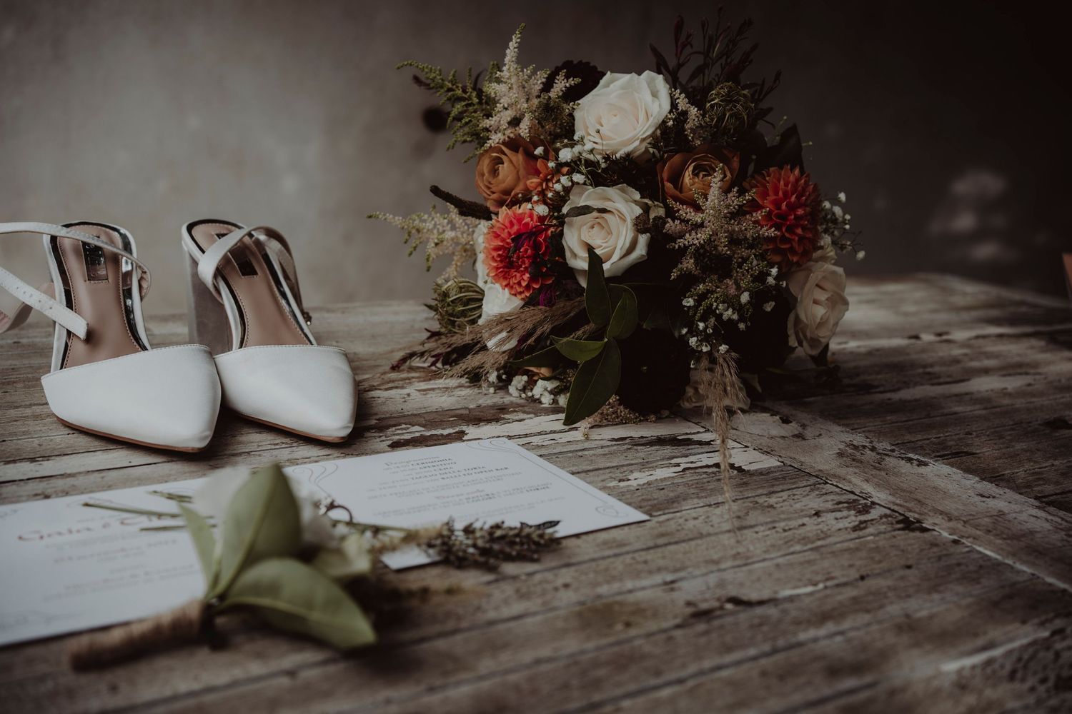 White bridal shoes and floral bouquet arranged with wedding invitations on weathered wooden surface.