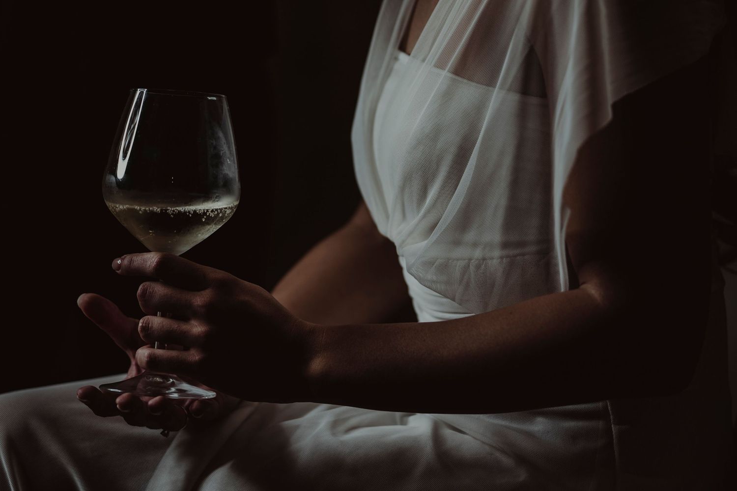 Person in white dress holding red wine glass against dark background.