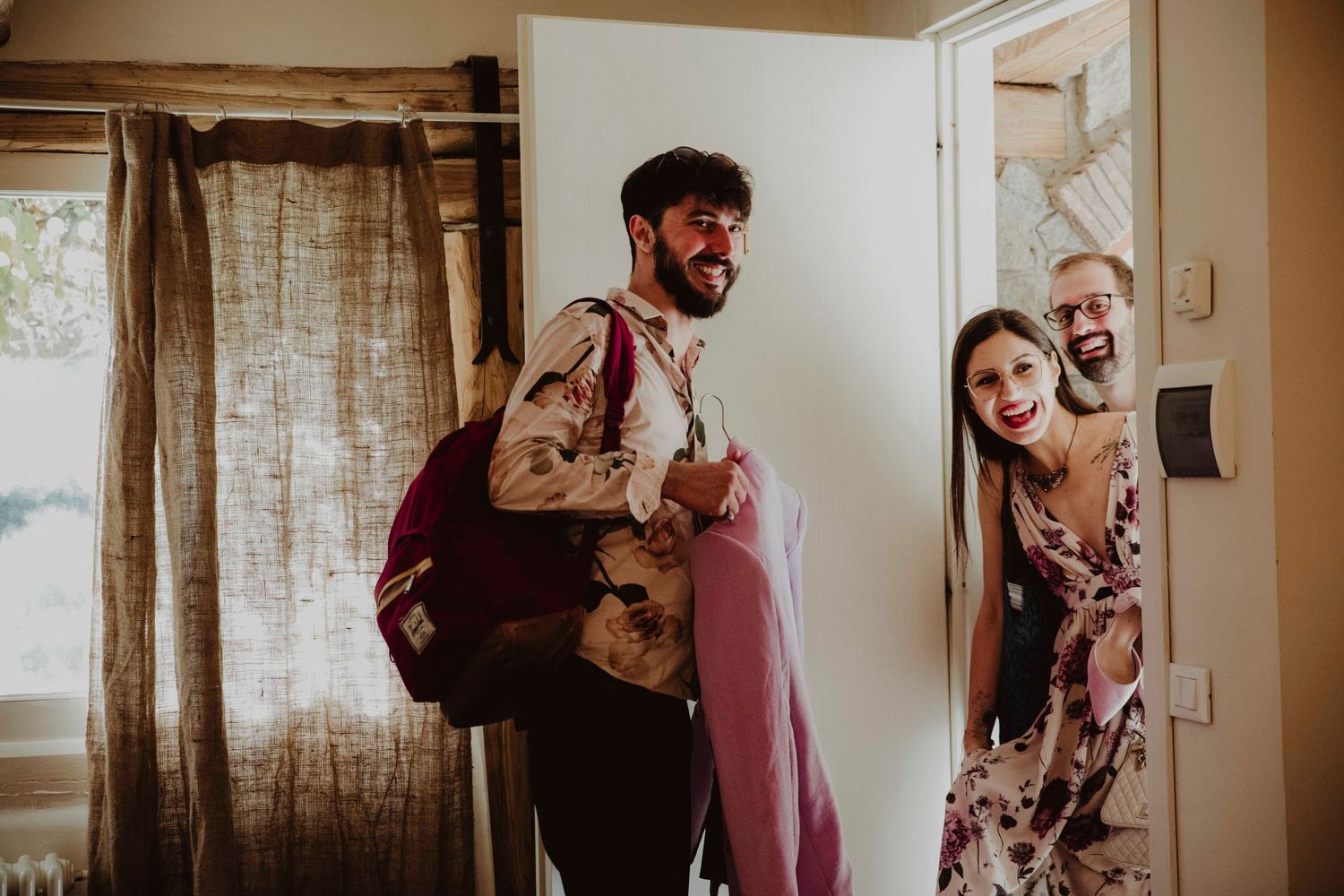 Wedding party members share a joyful moment in a rustic room with linen curtains.