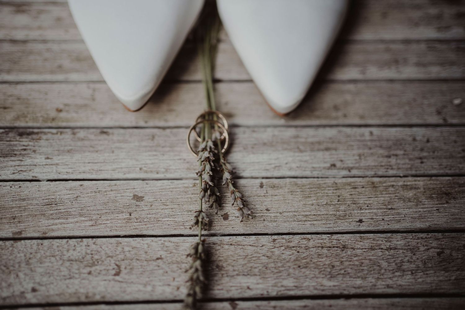 Close-up of white bridal shoes and dried flowers on weathered wooden floorboards.