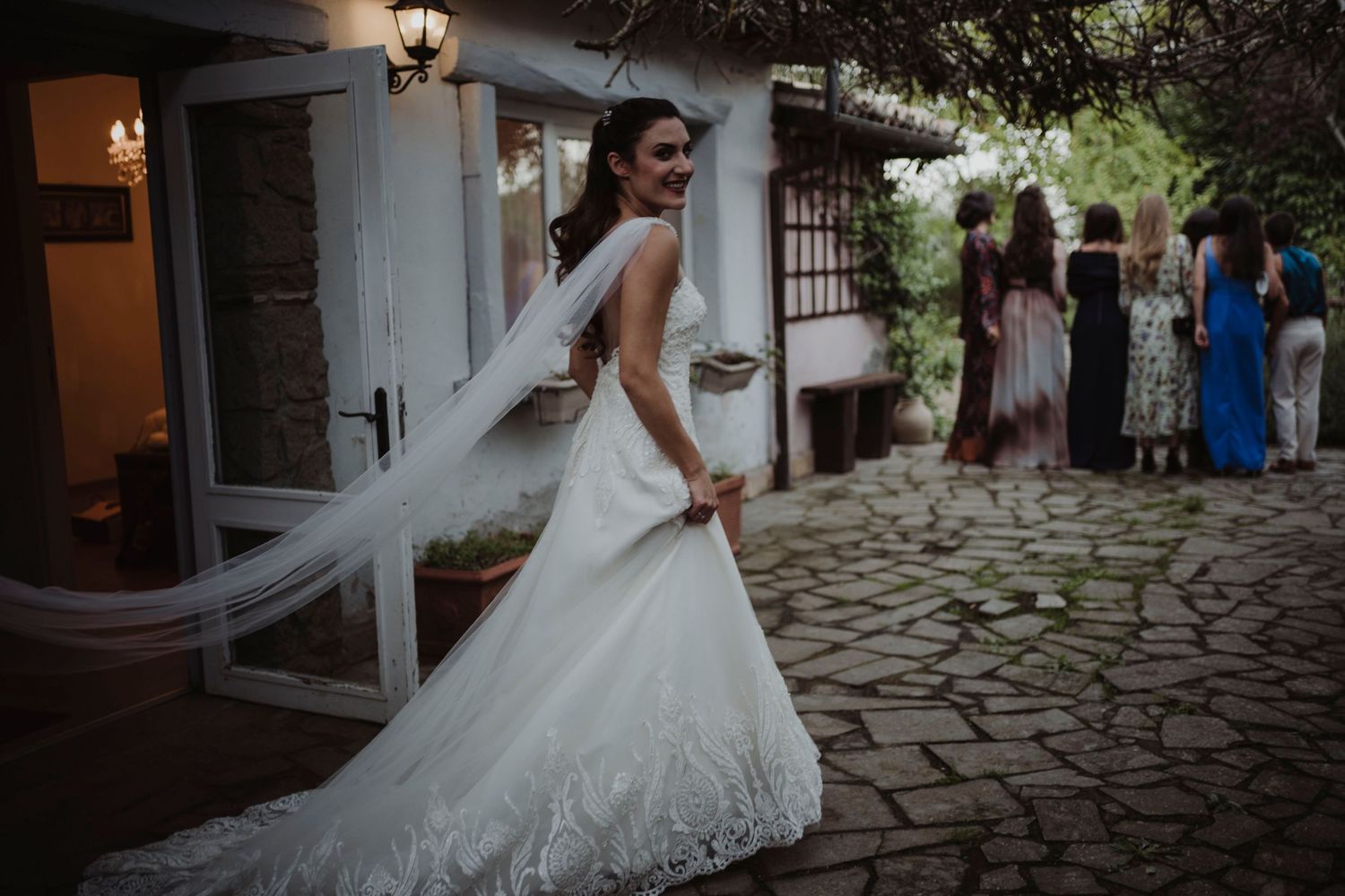 Bride in elegant lace wedding gown with flowing veil on stone pathway.