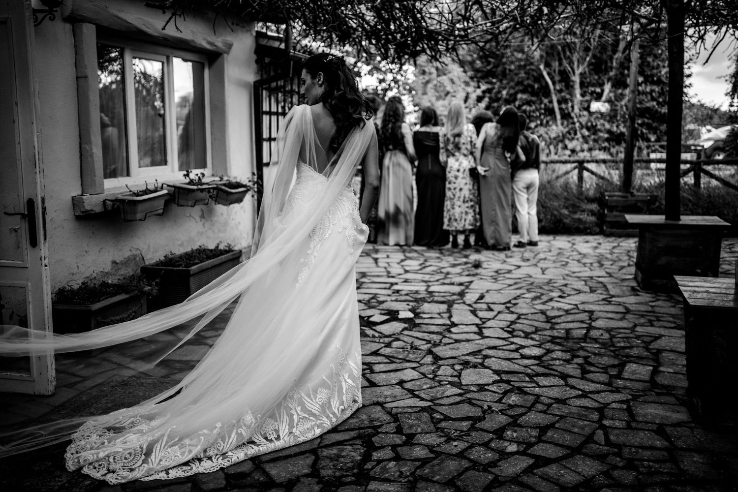Black and white photo of wedding celebration on cobblestone path near rustic building.