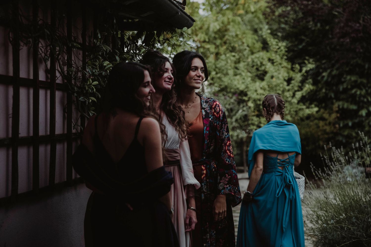 Wedding guests in colorful dresses sharing a moment outdoors near a rustic building.