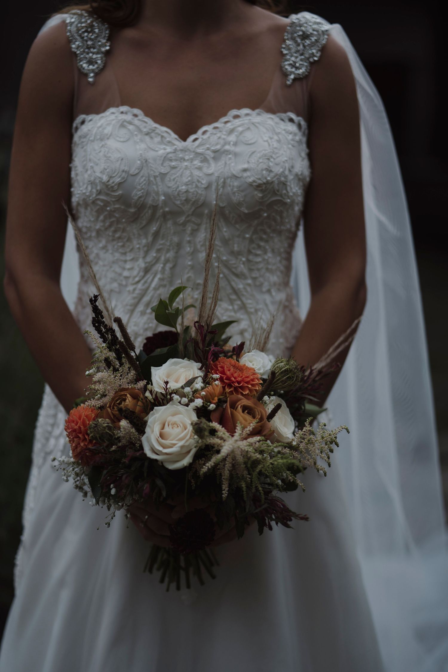 Close-up of a white lace wedding dress bodice and bouquet featuring roses and fall-toned flowers.